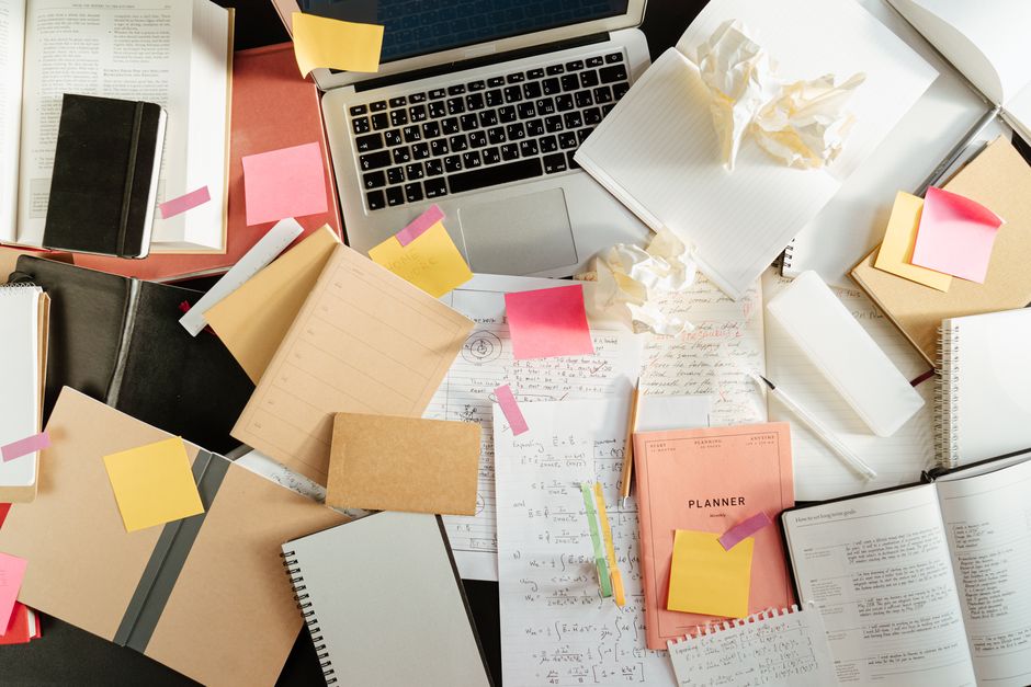 大量の書類、ノート、付箋、数式メモが積み重なった、情報が溢れたデスクの俯瞰写真